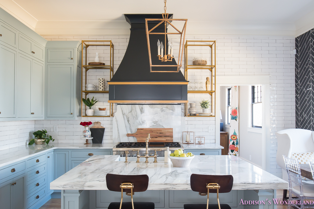 https://addisonswonderland.com/styling-open-kitchen-shelves/kitchen-white-marble-calcutta-gold-open-shelves-gold-black-vent-hood-blue-gray-cabinets-shaker-style-black-chevron-tile-subway-white-backsplash-decor-ideas-15-of-32/